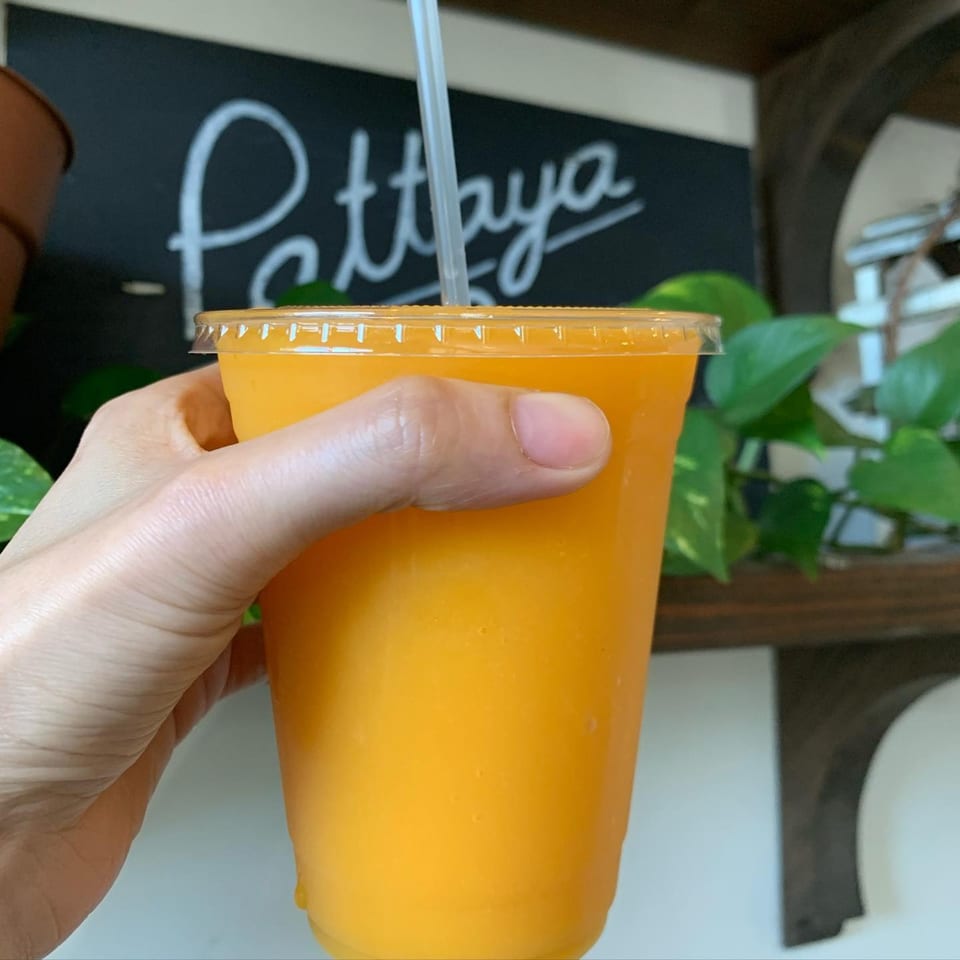 A hand holds a plastic cup of bright orange Thai iced tea with a straw, against a chalkboard.