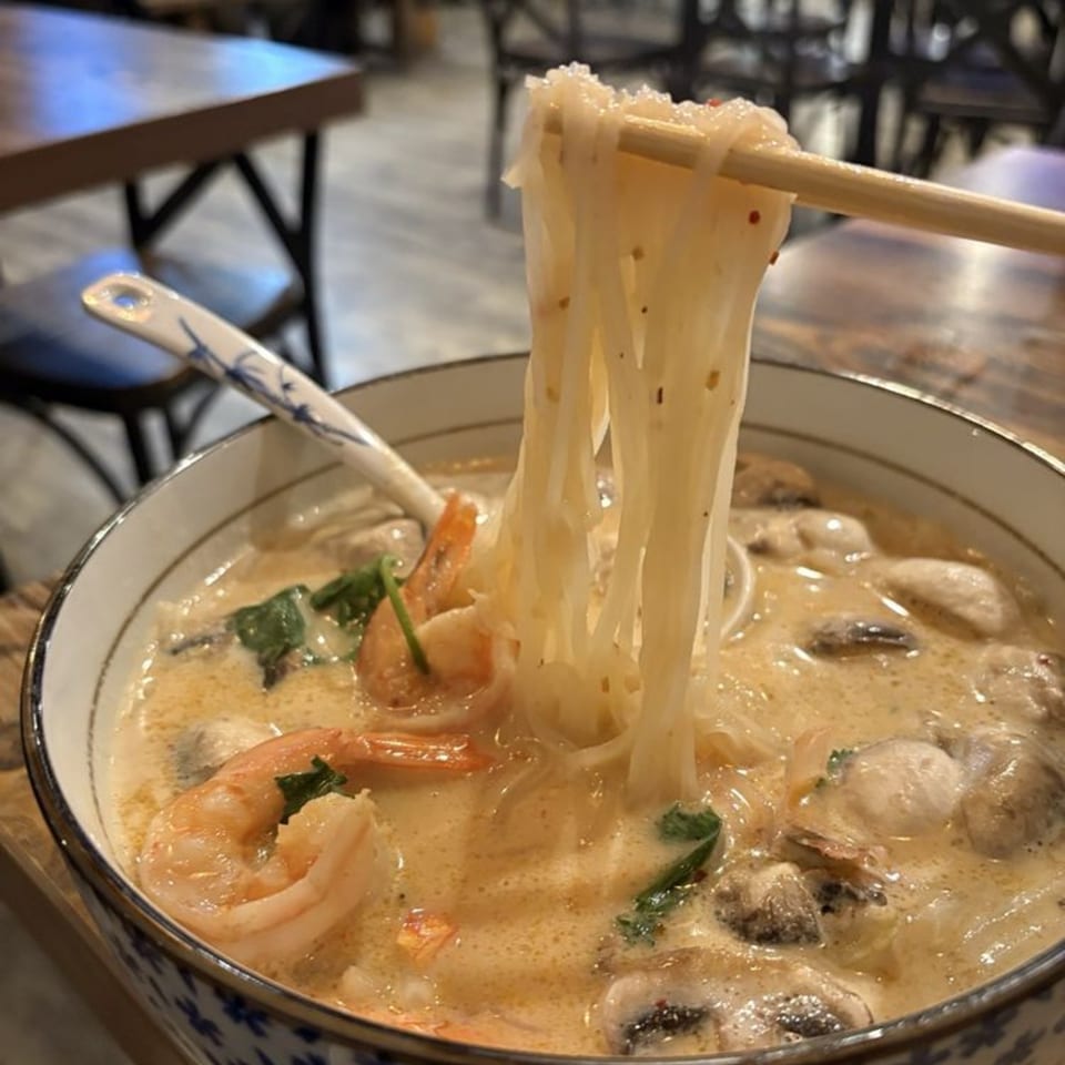 Chopsticks lifting noodles from a bowl of creamy Thai soup with shrimp and mushrooms.