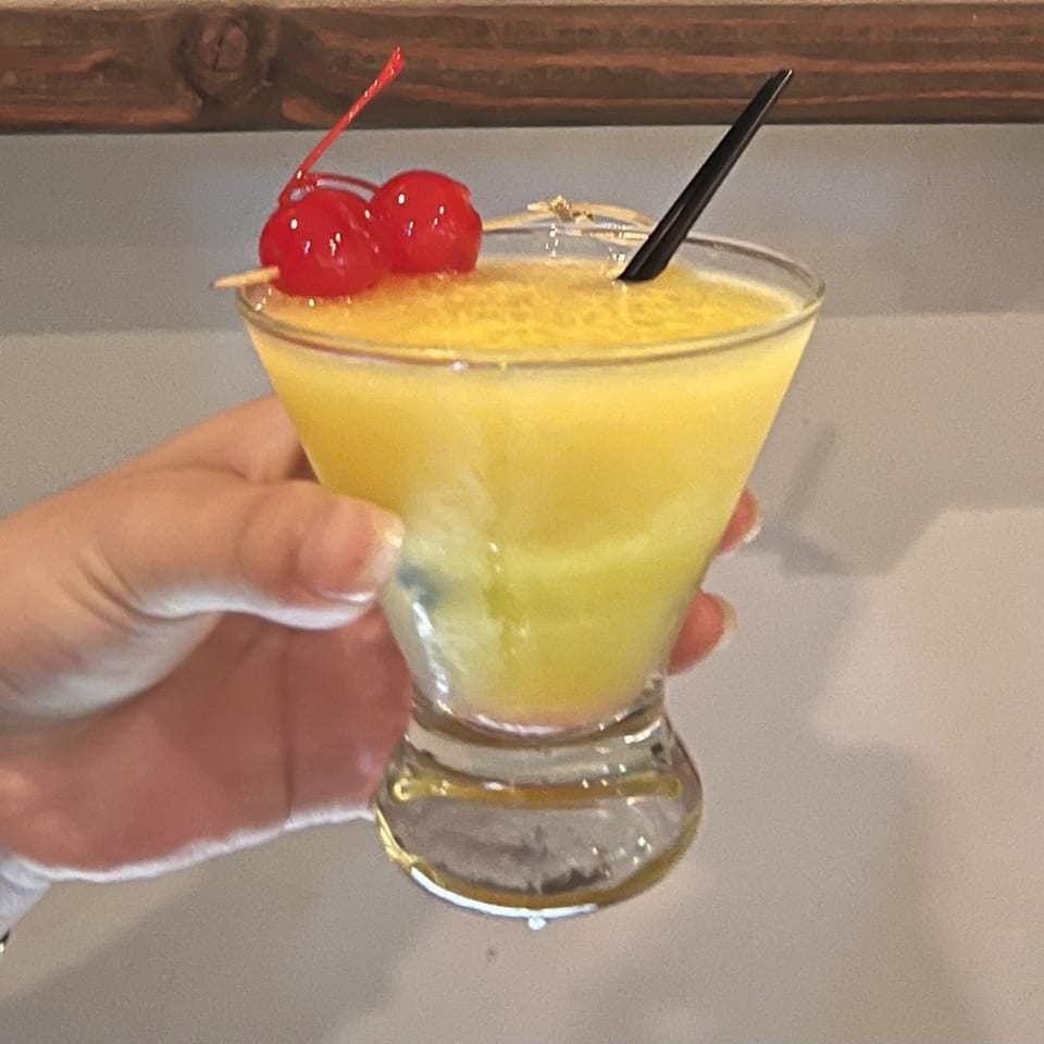 Hand holding a yellow cocktail garnished with two red cherries and a black straw.
