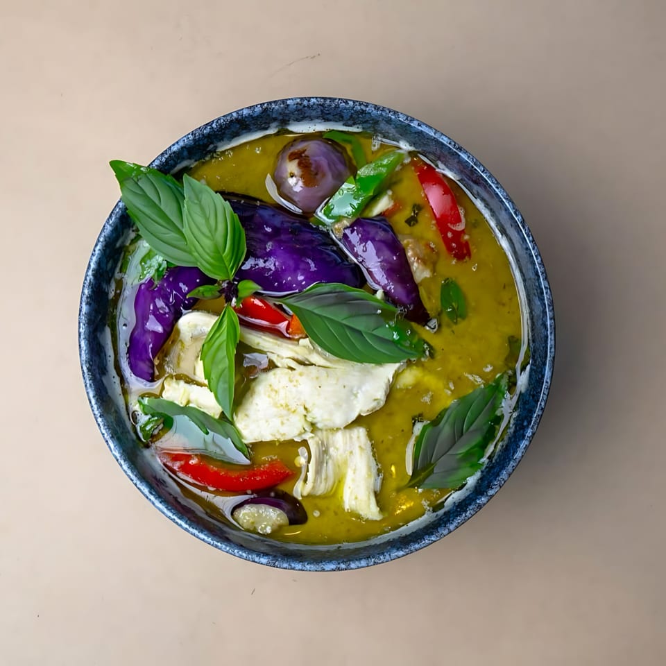 Overhead view of a bowl of green curry with chicken, purple eggplant, red chilies, and basil.