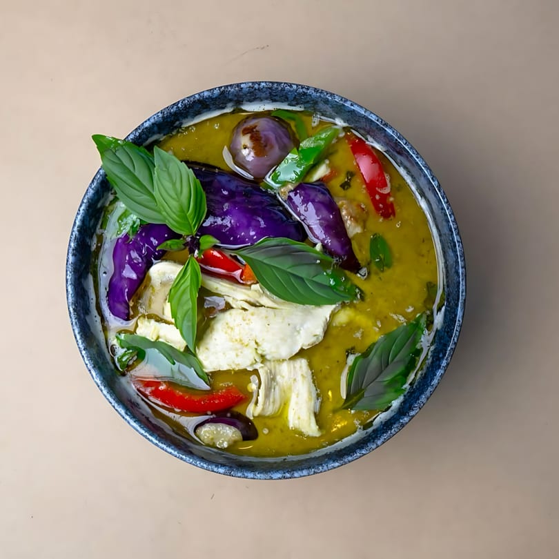 Overhead view of a bowl of green curry with chicken, purple eggplant, red chilies, and basil.